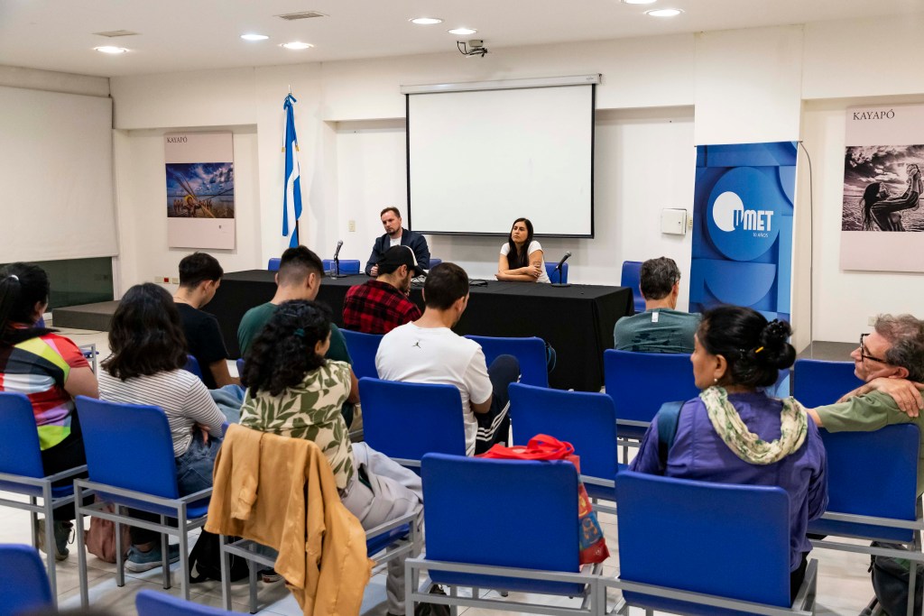 Mesa de debate en la Universidad Metropolitana para la Educación y el Trabajo (UMET), con dos expositores en el escenario y un público atento sentado en sillas azules.