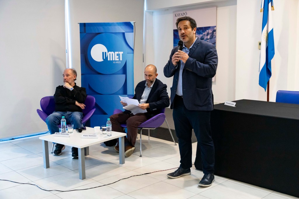 Un panel de discusión en la Universidad Metropolitana para la Educación y el Trabajo (UMET) con tres oradores, uno de ellos hablando al micrófono, mientras los otros dos escuchan. Bandera argentina en el fondo y logo de UMET visible.