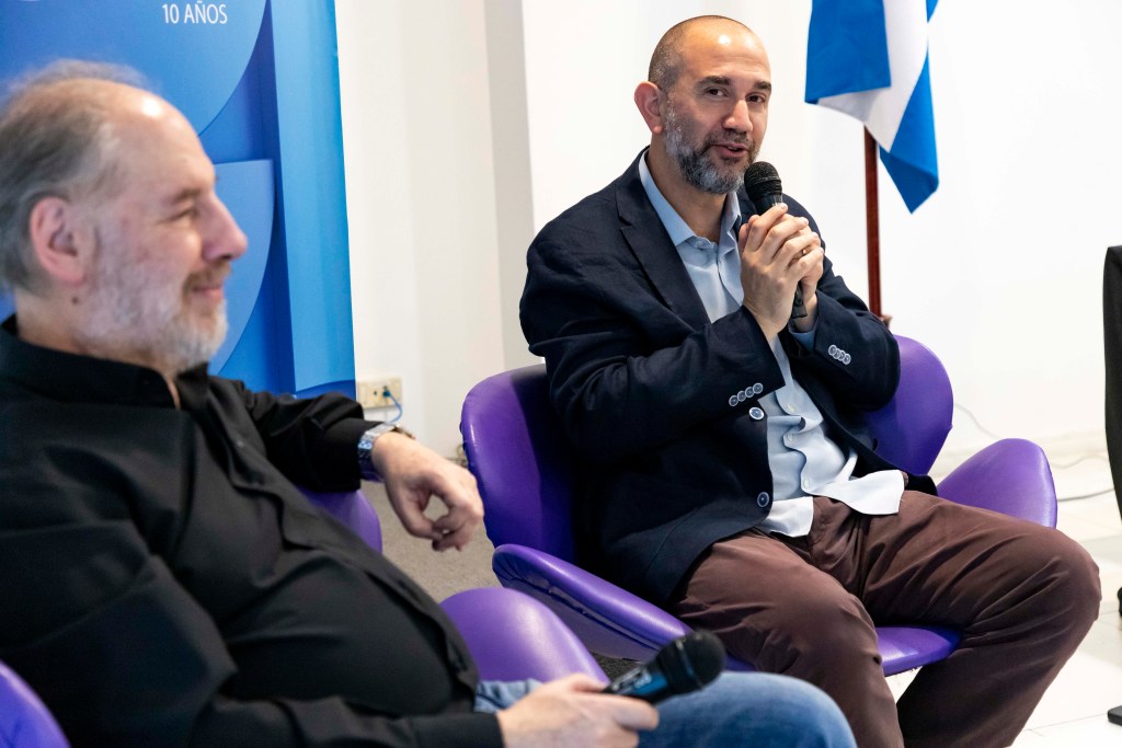 Dos hombres sentados en sillas moradas, uno hablando con un micrófono y el otro sonriendo, en un evento de la Universidad Metropolitana para la Educación y el Trabajo.