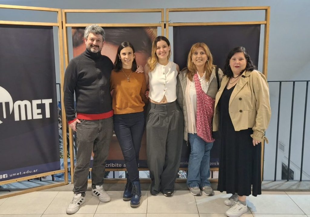 Un grupo de cinco personas posando frente a una pared con el logo de UMET. Todos sonríen y están vestidos de manera casual. Hay un ambiente amigable y profesional.