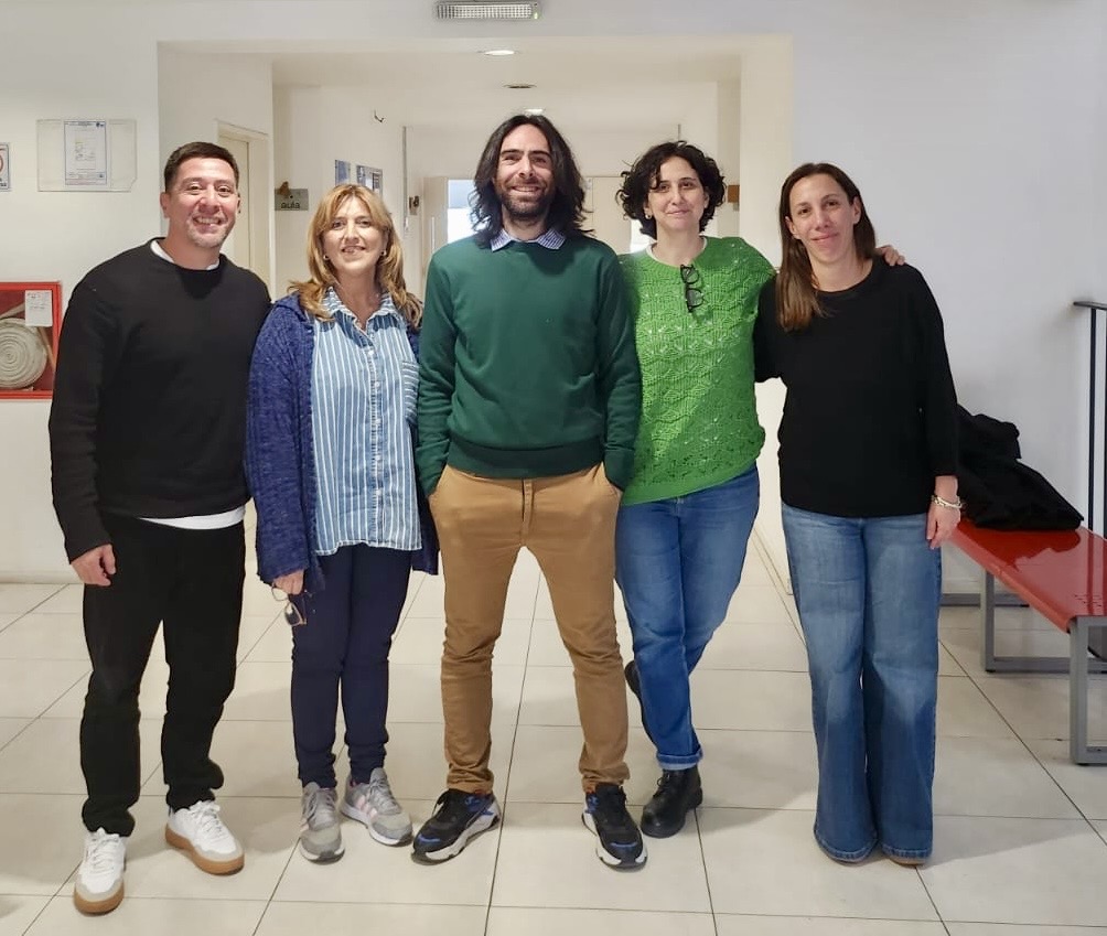 Cinco personas sonriendo en un recinto interior con paredes blancas y suelo de cerámica. Un hombre de pie en el centro viste un suéter verde y pantalones beige, rodeado por mujeres con diversas prendas, incluyendo una camisa a rayas y una camiseta negra.