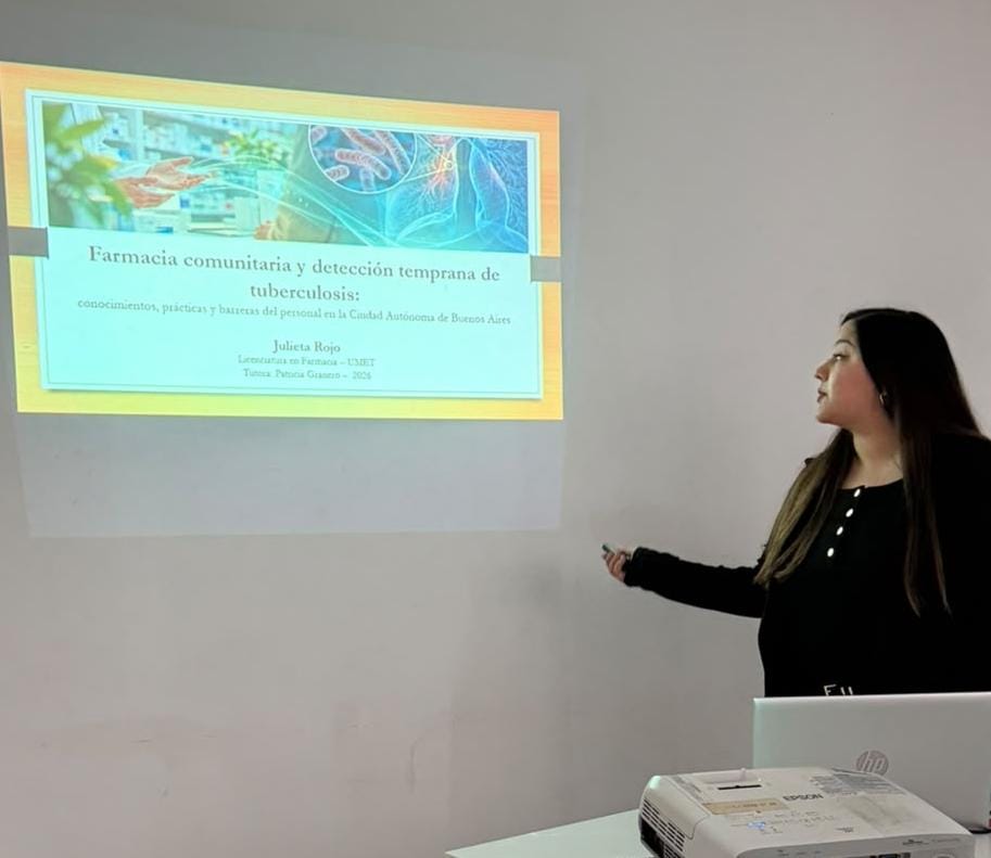 Una mujer presenta un tema sobre 'Farmacia comunitaria y detección temprana de tuberculosis' en una pantalla, con un proyector y una computadora portátil visible.